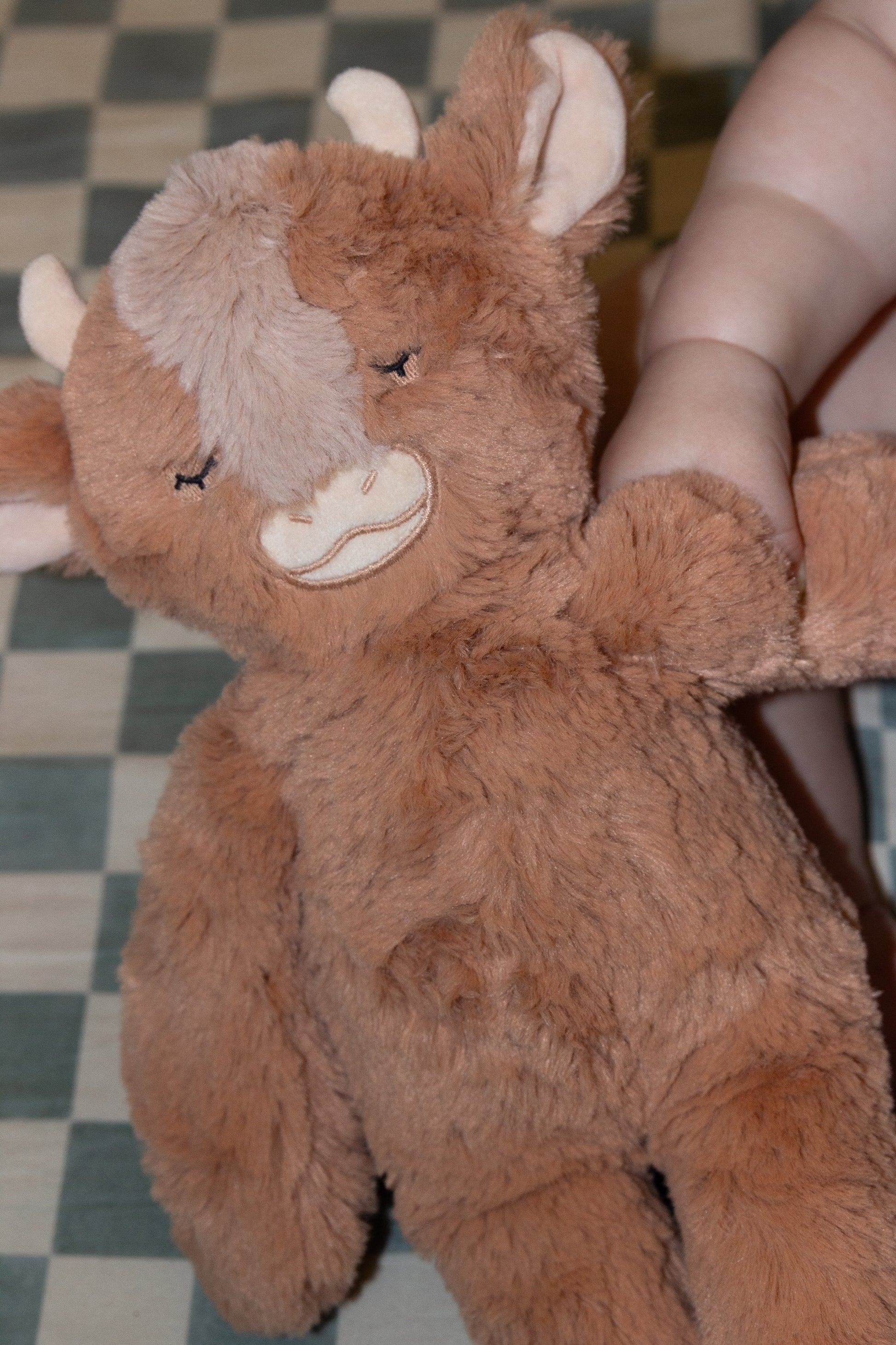 Brown plush toy resembling a moose held by a hand on a checkered floor.
