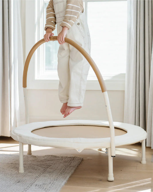Person using a small trampoline in a room with light-colored curtains and wooden floor.