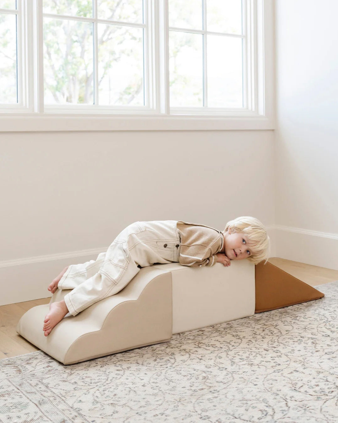 Child lying on a white wedge cushion in a bright room with large windows.